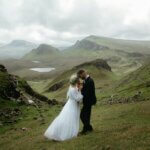A bride and groom standing on a grassy hill