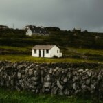 Small white house on a green hill with stone wall.